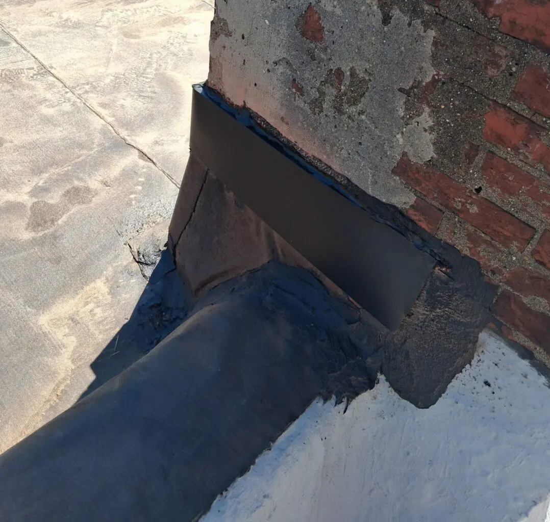 A black pipe connected to a brick wall with flashing and sealant.