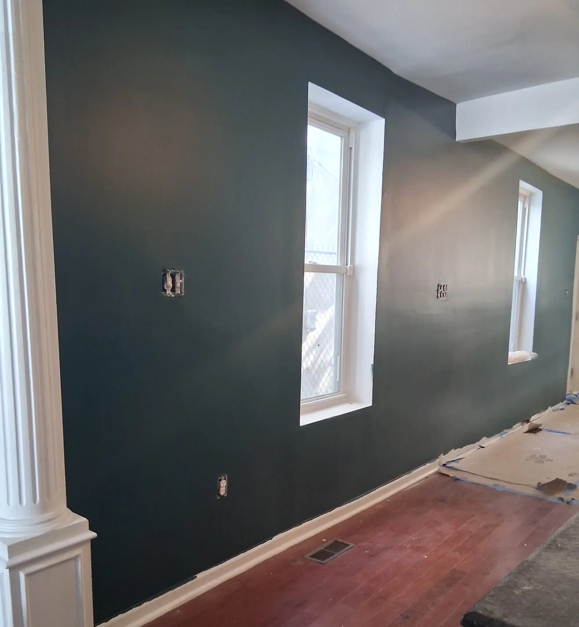 A dark-painted wall with two windows and white trim in a room under renovation.