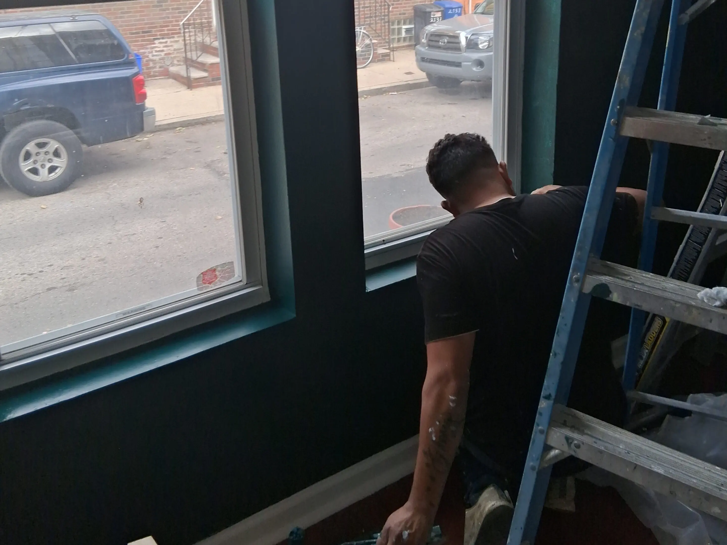A man painting a dark blue wall near a window with a ladder nearby.